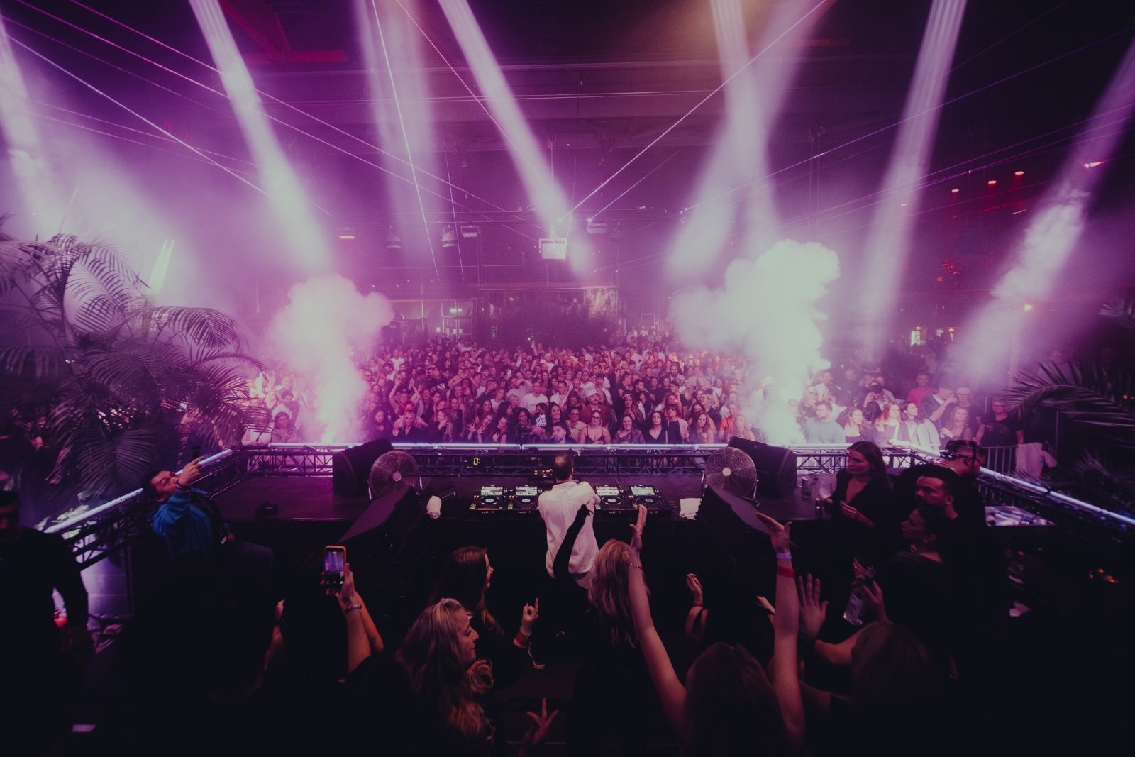 Packed dancefloor seen from behind the DJ booth, light beams and smoke over the crowd
